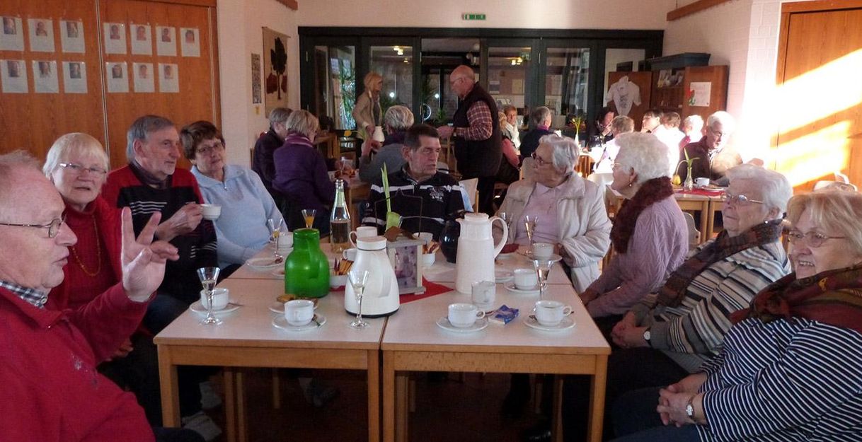 A group of elderly people is sitting at tables in a bright room. They are happily chatting over coffee and cake.