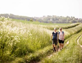 Zwei Pilgerinnen sind auf einem Feldweg zwischen hohem Gras zu sehen.