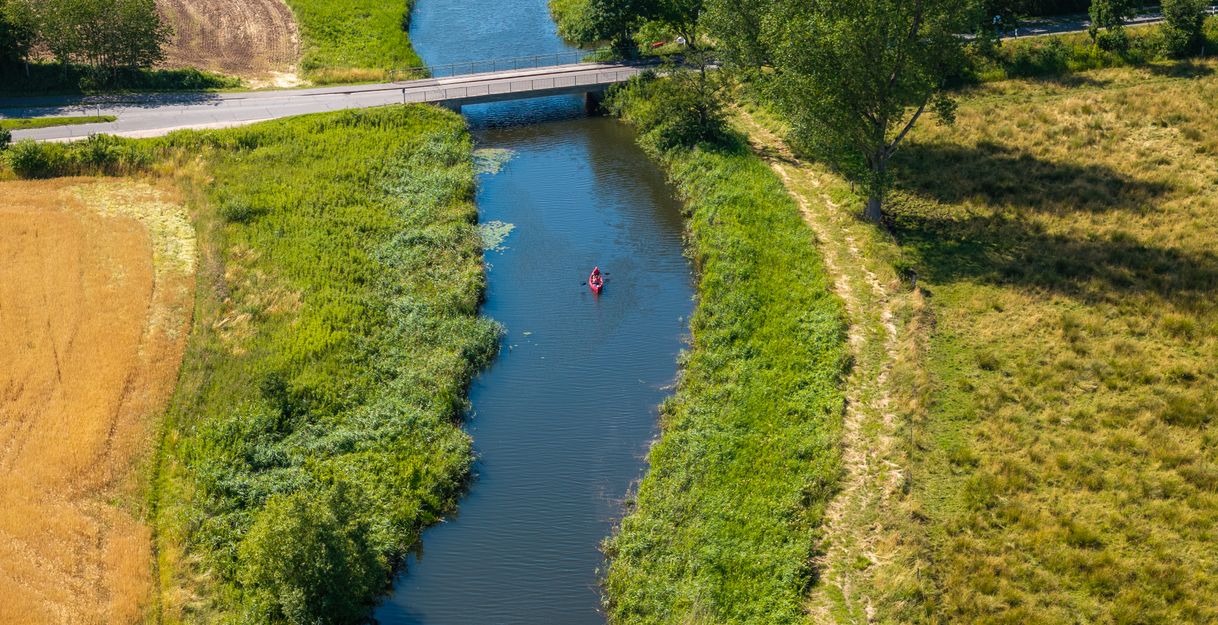 Luftaufnahme eines schmalen Flusses mit Brücke in Schleswig-Holstein – grüne Uferlandschaft und ein Kanufahrer in ruhiger Naturidylle.