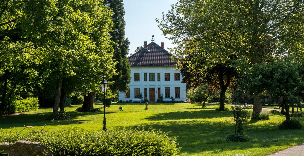 Ein weißes Herrenhaus steht inmitten eines grünen Parks mit Bäumen und einem gepflegten Rasen.