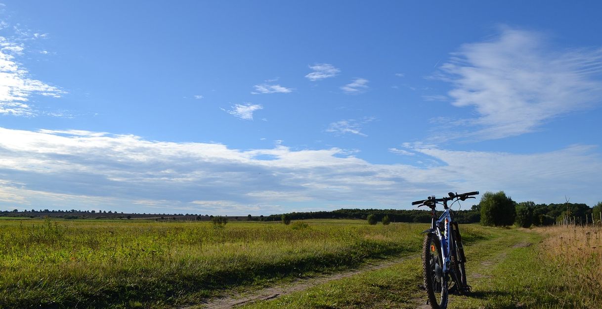 Ein Fahrrad steht auf einem Feldweg. Es ist blauer Himmel mit Wolken in der Ferne zu sehen und Grüne Wiese säumt den unbefiestigten Weg.