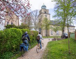 Zwei Radfahrer fahren auf einem Weg zur Vicelinkirche in Neumünster, umgeben von blühenden Bäumen und grüner Vegetation.