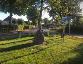 Es ist ein Gedenkstein auf einer Wiese im Streiflicht der warmen Nachmittagssonne zu sehen.