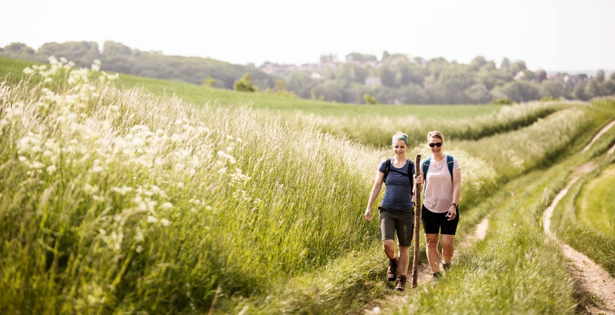 Zwei Pilgerinnen sind auf einem Feldweg zwischen hohem Gras zu sehen.